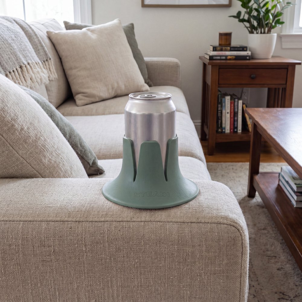 A can standing in a sage Steadi on the linen arm of a sofa, a side table stacked with books behind it.