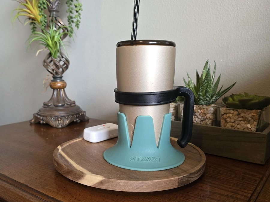 A champagne-gold tumbler with a black-and-white striped straw in a turquoise STEADI base, standing on a round wooden serving board on a dark wood side table with a lamp, a small remote and potted succulents